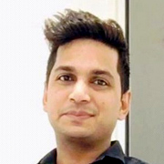 A close-up photo of a smiling man with short dark hair, dressed in a black shirt, against a light-colored background.