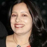 A woman smiling brightly, with dark hair and a necklace, dressed in a black top with red patterns.