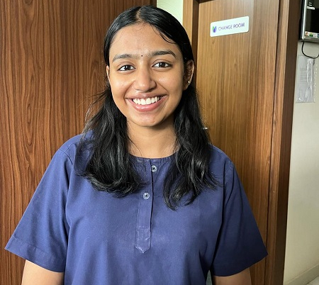A portrait of a young woman wearing blue scrubs, smiling warmly in a clinical setting with a "Change Room" sign visible in the background.