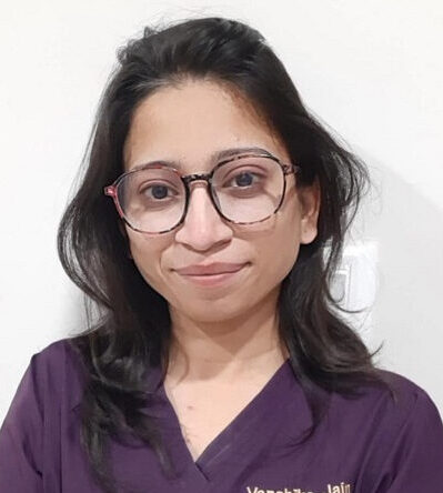 A portrait of a female healthcare professional wearing purple scrubs and glasses, smiling warmly in a clinical setting.