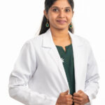 A professional portrait of a female doctor wearing a white lab coat, with a green outfit underneath and a confident smile.