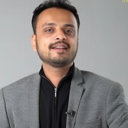 A professional man wearing a grey jacket, black shirt, and speaking directly to the camera with a friendly smile.