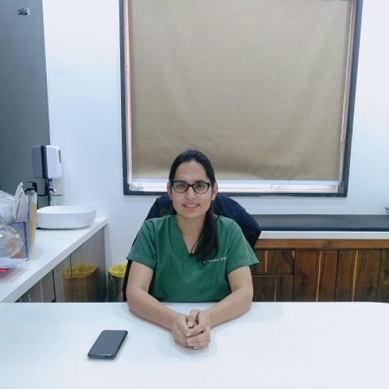 A portrait of a female professional sitting at a desk in a medical setting, wearing green scrubs, with glasses and a calm expression.