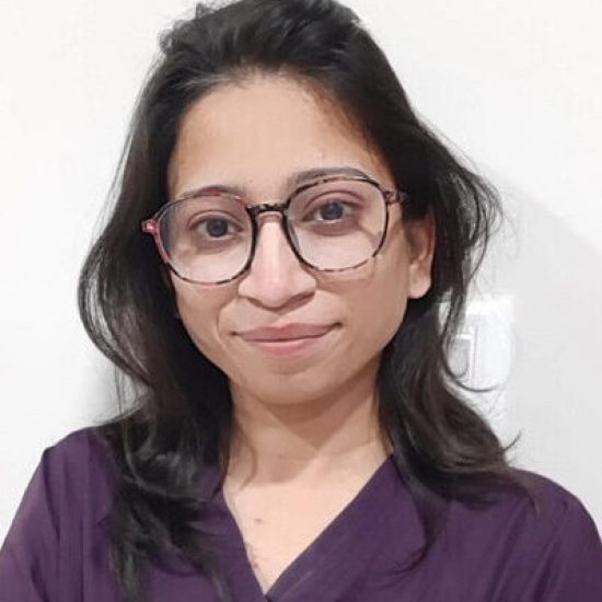 A portrait of a female healthcare professional wearing purple scrubs and glasses, smiling warmly in a clinical setting.