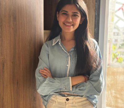 A portrait of a young woman smiling and standing with her arms crossed, wearing a denim shirt and beige pants in a bright indoor setting.