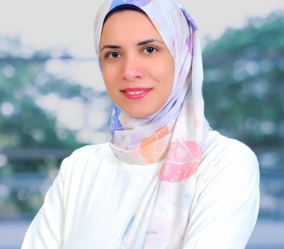 A portrait of a female healthcare professional wearing a white blouse and a light-colored hijab, smiling warmly in a serene outdoor setting.