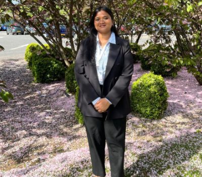 A portrait of a young woman standing in a park or garden, dressed in a formal black blazer, white blouse, and black trousers, with a pleasant smile and a relaxed posture.